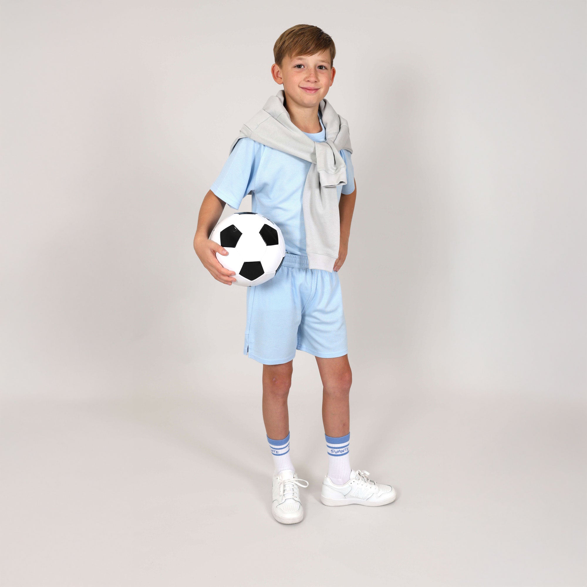 Boy aged ten holding a football under his arm with a regenerative cotton hoody over his shoulders and wearing sky blue sports shorts and sports t-shirt made from toxin free, natural materials