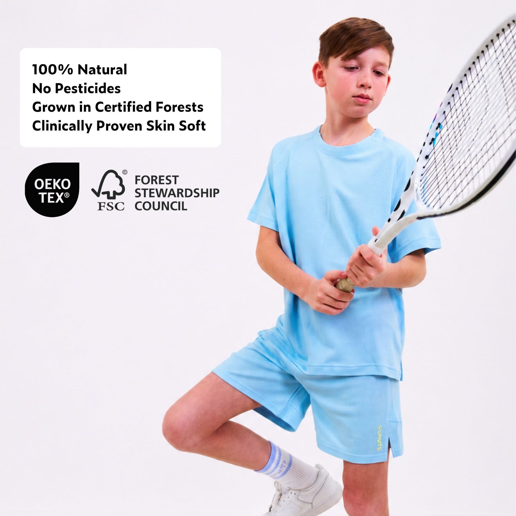 Boy in light blue tencel sports shorts and sports t-shirt holding a tennis racket with certification logos on a white background