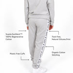 A girl shown from the back wearing Svante organic cotton light grey sweatpants with white sneakers, on a white studio background. Text callouts point to features of the sweatpants, including Svante Earth knit 100% regenerative cotton, toxin free natural silicone print down the side, plastic free cuffs, and organic cotton stitching.