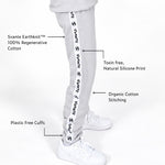 Svante Regenerative Cotton Light grey sweatpants shown on a model from the waist down, featuring a prominent white side stripe with repeated black 'SVANTE' text. Callout lines and text describe product features including Svante Earthknit 100% regenerative cotton, toxin-free natural silicone print, organic cotton stitching, and plastic-free cuffs.