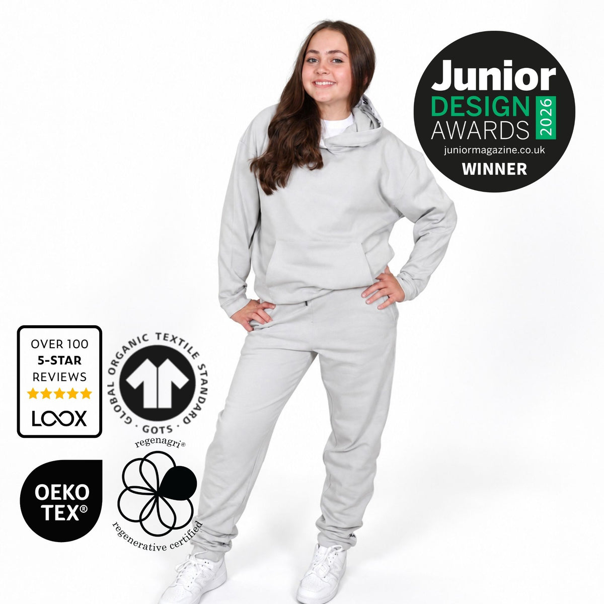 Girl modelling a Svante regenerative cotton light grey two-piece tracksuit, featuring a matching hoodie and sweatpants, set against a plain white background. The image includes several badges on the left and right, including 'Junior Design Awards 2026 Winner' and certification logos for LCOOX and OEKO-TEX. 