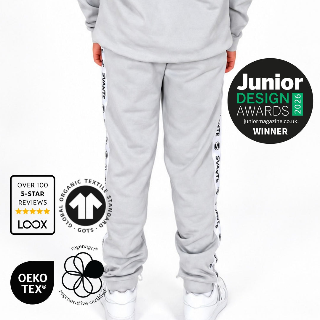Boy shown from the back wearing a Svante organic cotton hooded sweatshirt and matching organic jogger pants with white sneakers. The image includes award and certification badges on the left and right, including Junior Design Awards 2026 Winner, LOOX, OEKO-TEX, and GOTS logos, on a plain white background. 