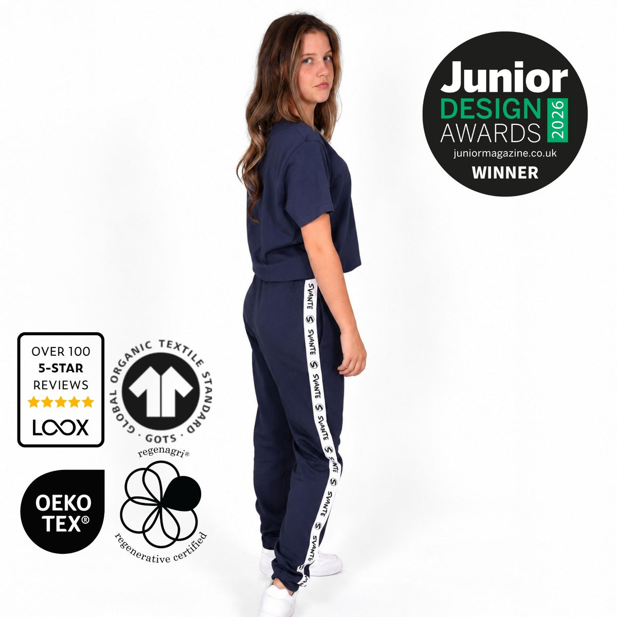 A girl modelling a Svante organic cotton navy blue two-piece tracksuit with white text details on the pants, shown against a white background with several certification and Junior Design Awards award badges. 