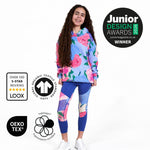A young girl stands against a white background wearing a Svante regenerative cotton light blue floral hoodie and matching blue leggings with a colourful floral print. The image includes award badges and certification logos: Junior Design Awards 2026 Winner, LOOX over 100 5-star reviews, OEKO-TEX, and GOTS organic textile standard.