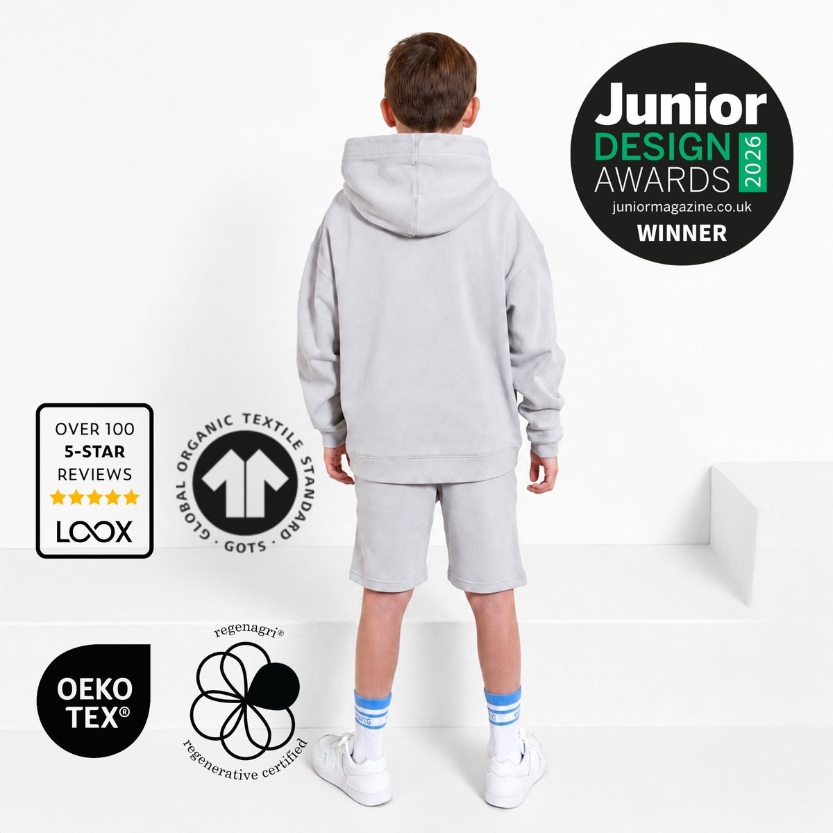 Boy with back to camera wearing a Svante grey organic cotton hoodie and grey organic cotton jersey shorts standing on a white platform. The image includes award and certification badges: a Junior Design Awards 2026 winner badge, LOOX over 100 5-star reviews badge, OEKO-TEX badge, and a Global Organic Textile Standard badge. 
