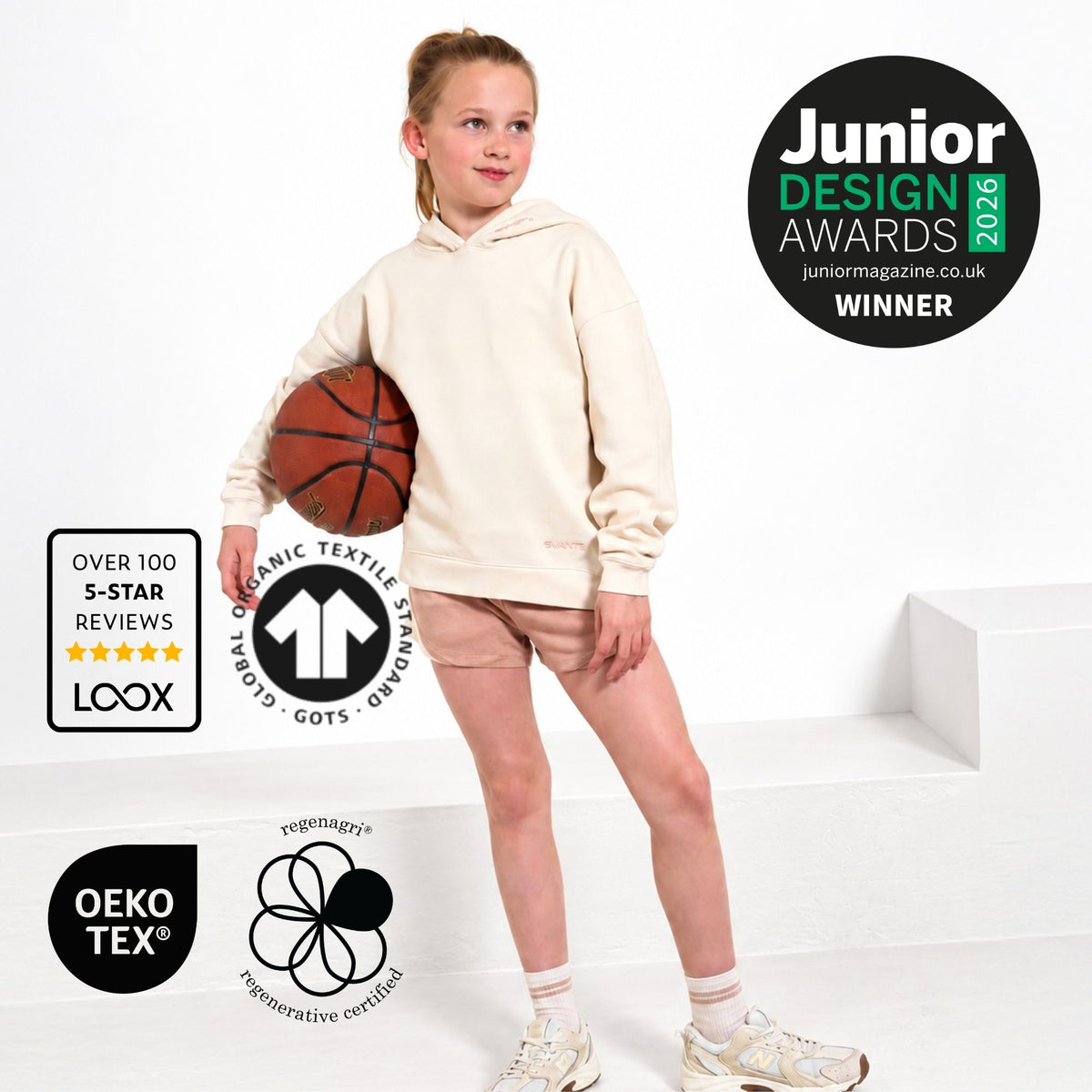 A young girl stands on white steps wearing a Svante regenerative cotton cream-coloured hoodie and polyester free sports shorts, holding a basketball. The image includes several certification and award badges, including 'Junior Design Awards 2026 Winner' from Junior Magazine and 'Over 100 5-Star Reviews' from LOOX.