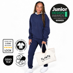 A young girl modelling a Svante regenerative cotton navy blue two-piece tracksuit, holding a cream canvas tote bag with 'SVANTE natural movement.' printed on it. The image includes several certification and award badges on the left and a 'Junior Design Awards 2026 Winner' badge on the right, all against a solid white background. 