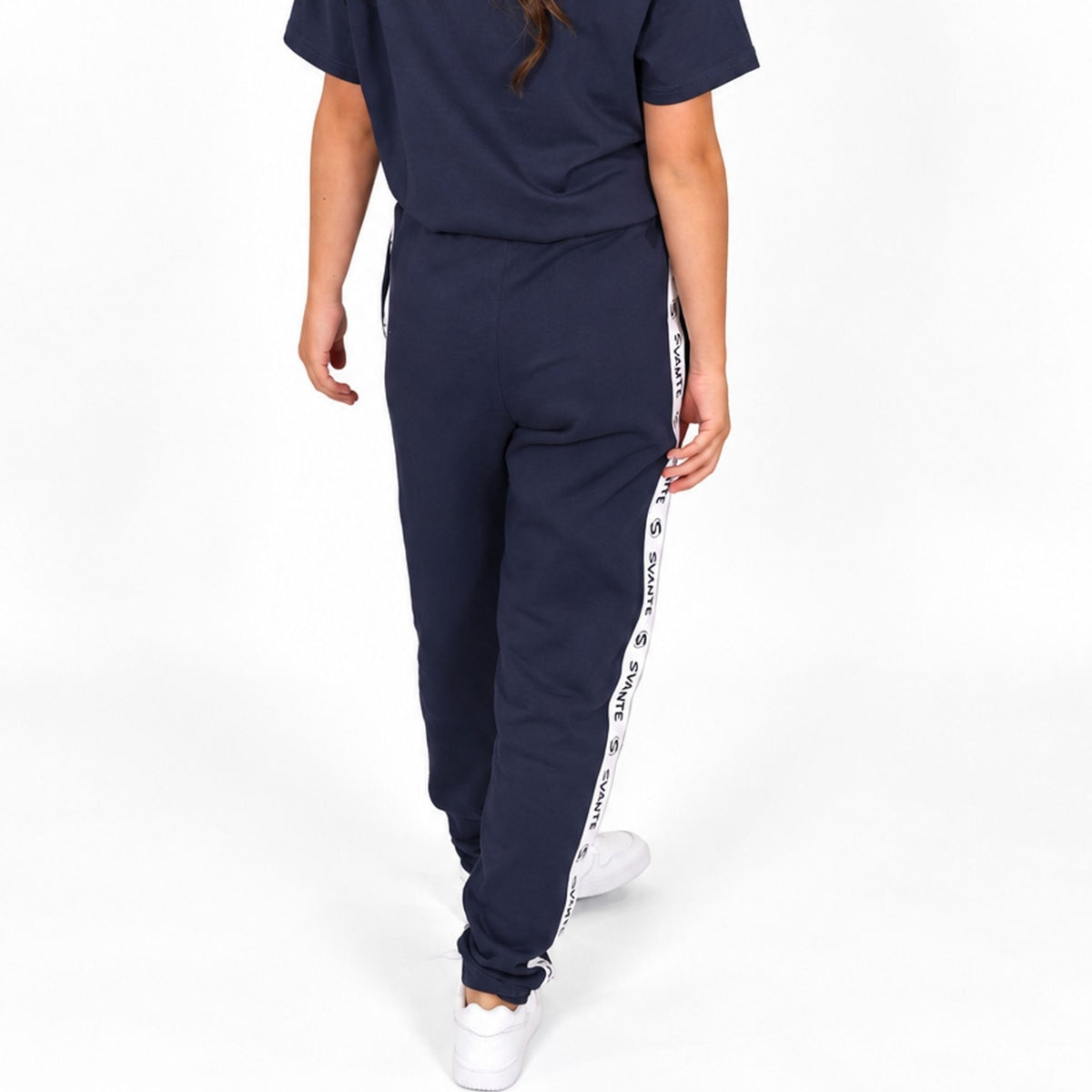 A girl wearing Svante regenerative and organic cotton navy blue sweatpants with a white side stripe featuring black text, paired with a matching navy t-shirt and white sneakers, shown from the back against a white studio background.