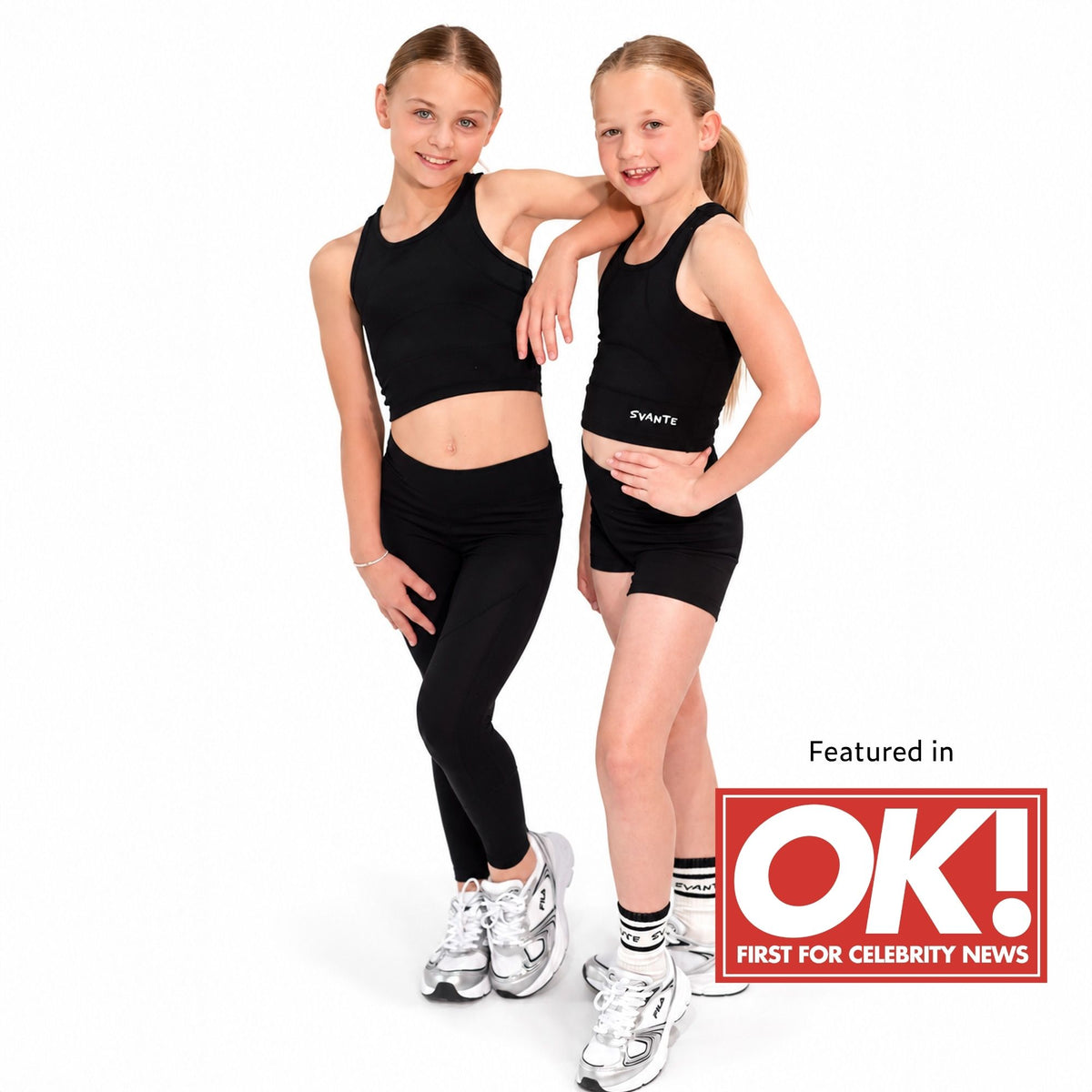 Two young girls modelling Svante polyester free black activewear sets, one in a sports crop top and leggings and the other in a sports crop top and shorts, standing against a white background. A red 'Featured in OK! First For Celebrity News' logo is visible in the bottom right corner.