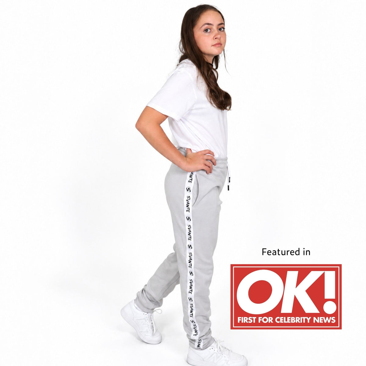 A girl wearing Svante organic cotton light grey sweatpants with black text down the side, a white t-shirt, and white sneakers, standing against a white background. A red and white 'Featured in OK! FIRST FOR CELEBRITY NEWS' badge is in the bottom right corner. 