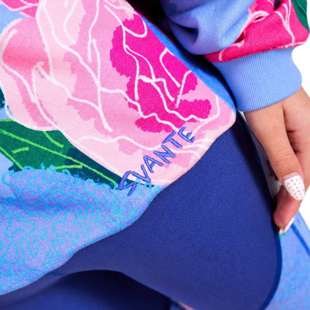 Close-up of a girl wearing a colourful Svante organic cotton floral sweatshirt and blue leggings, with a hand visible at the side. The sweatshirt features a pink and light blue floral pattern with the word 'SVANTE' embroidered in blue.