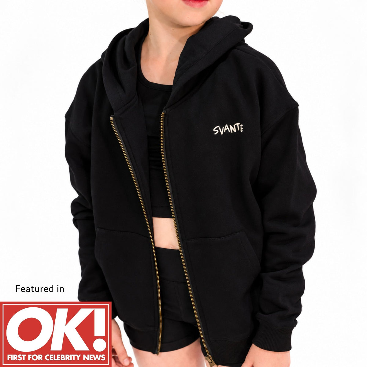 A girl wearing a svante organic cotton black zip-up hoodie with a gold zipper and a small white 'SVANTE' logo on the chest, shown against a plain white background with a red 'Featured in OK! First For Celebrity News' badge in the lower left.
