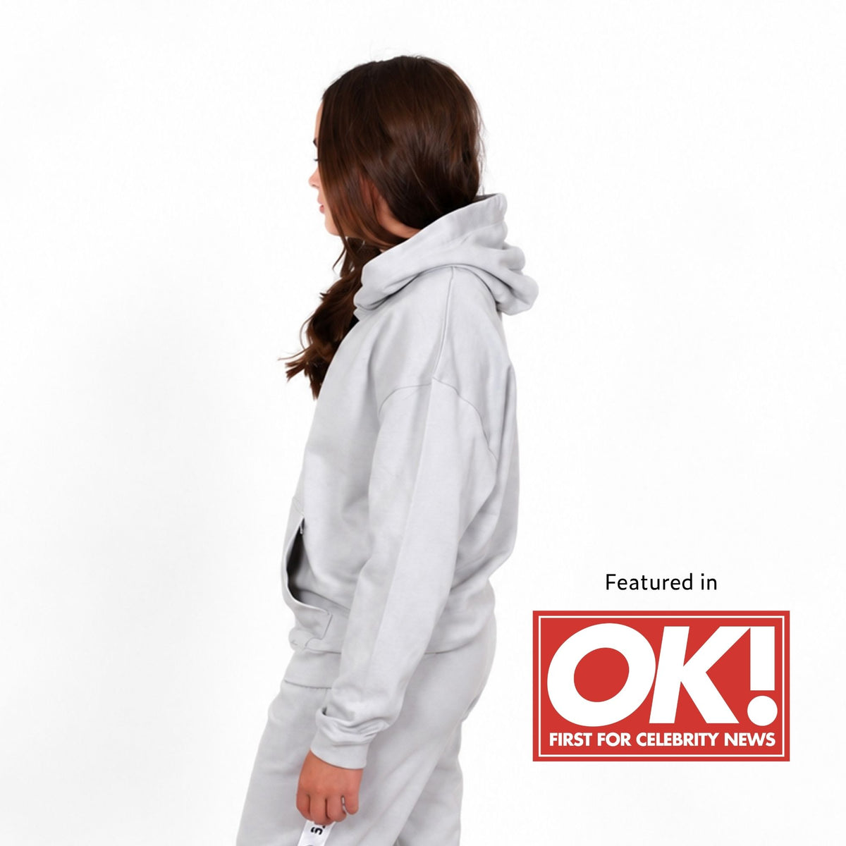 Teenage girl shown from the side wearing a svante organic cotton light grey hooded sweatshirt and matching sweatpants against a white background, with a 'Featured in OK! FIRST FOR CELEBRITY NEWS' graphic overlay in the bottom right corner. 