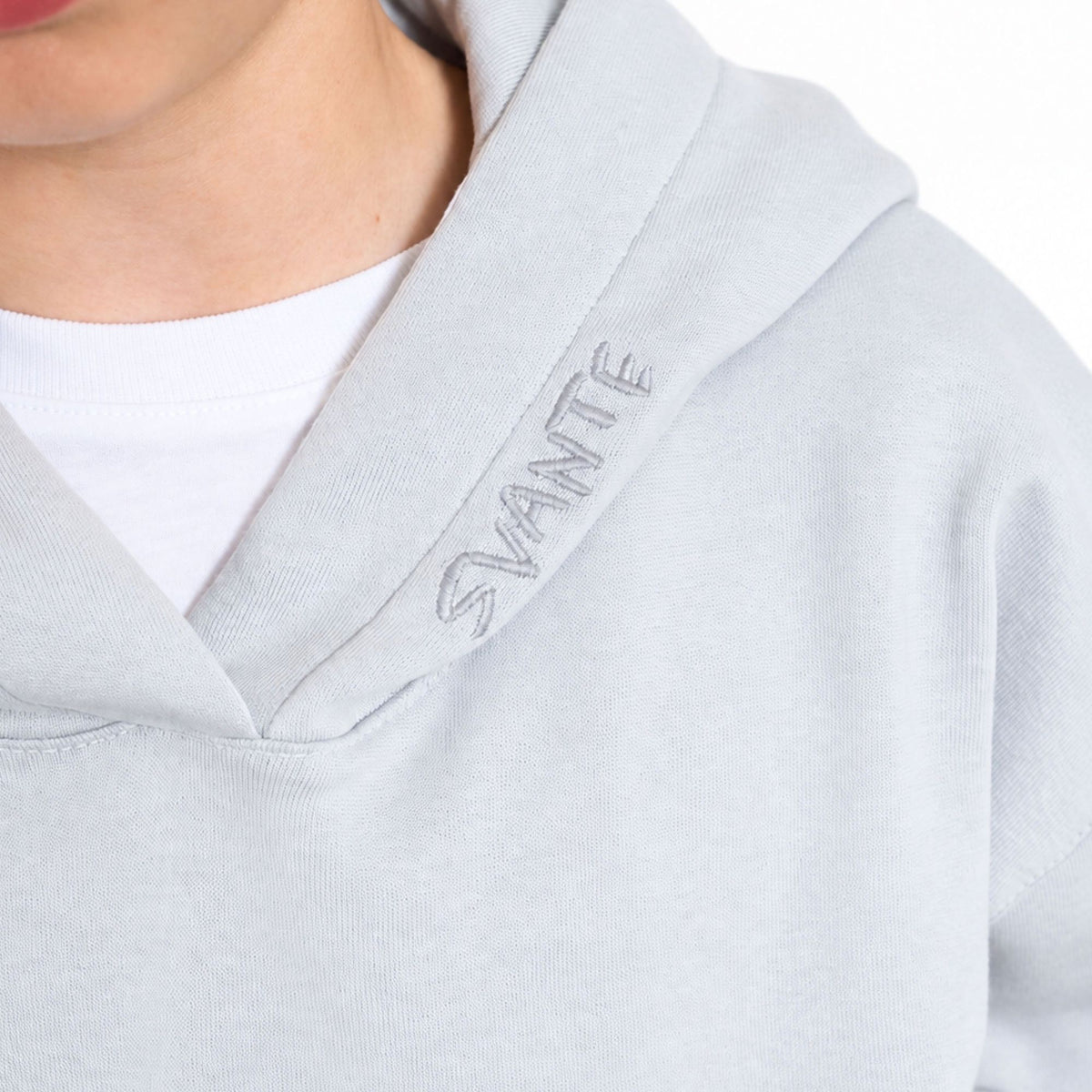 Close-up of a boy wearing a light grey regenerative cotton hoodie with 'SVANTE' embroidered vertically on the hood, layered over a white t-shirt. 