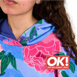 Close-up of a girl wearing a colourful Svante organic cotton floral hoodie with pink flowers, green leaves, and a blue base. A red and white 'Featured in OK! First For Celebrity News' badge is overlaid on the lower right.