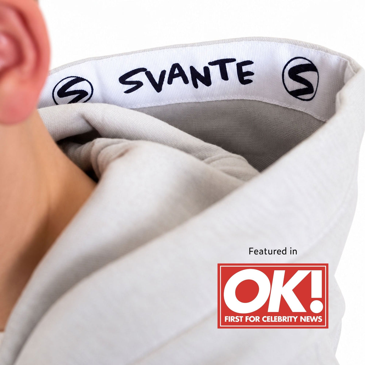 Close-up of a boy wearing a light grey organic cotton hoodie with a white inner band printed with the brand name 'SVANTE' and black 'S' logos. A red 'Featured in OK! First For Celebrity News' badge appears in the lower right. 