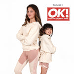 A teenage girl and a young girl wearing matching Svante organic cotton cream-coloured hoodies and light pink leggings, posed back-to-back against a white background. A red 'Featured in OK! FIRST FOR CELEBRITY NEWS' badge is in the top right corner.