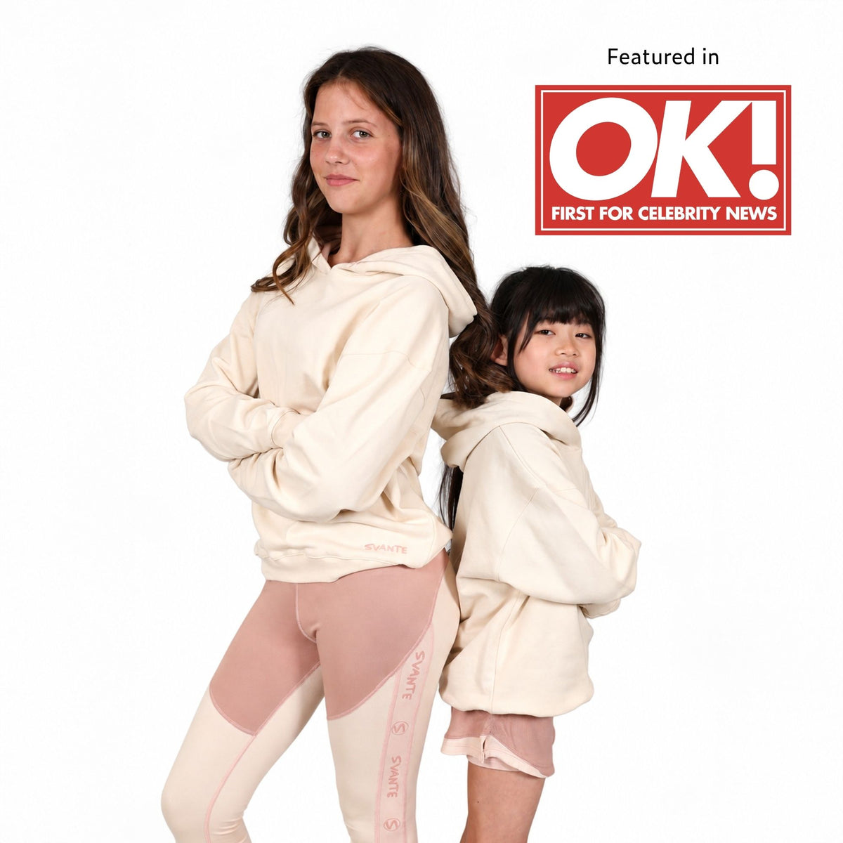A teenage girl and a young girl wearing matching Svante organic cotton cream-coloured hoodies and light pink leggings, posed back-to-back against a white background. A red 'Featured in OK! FIRST FOR CELEBRITY NEWS' badge is in the top right corner.