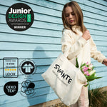 A child wearing a cream Svante sweatshirt holds a cream Svante organic cotton canvas tote bag printed with the brand name 'SVANTE' and 'natural movement' in black text, standing against a light blue wooden wall and holding pink tulips. Multiple certification and award badges are overlaid on the left side of the image.