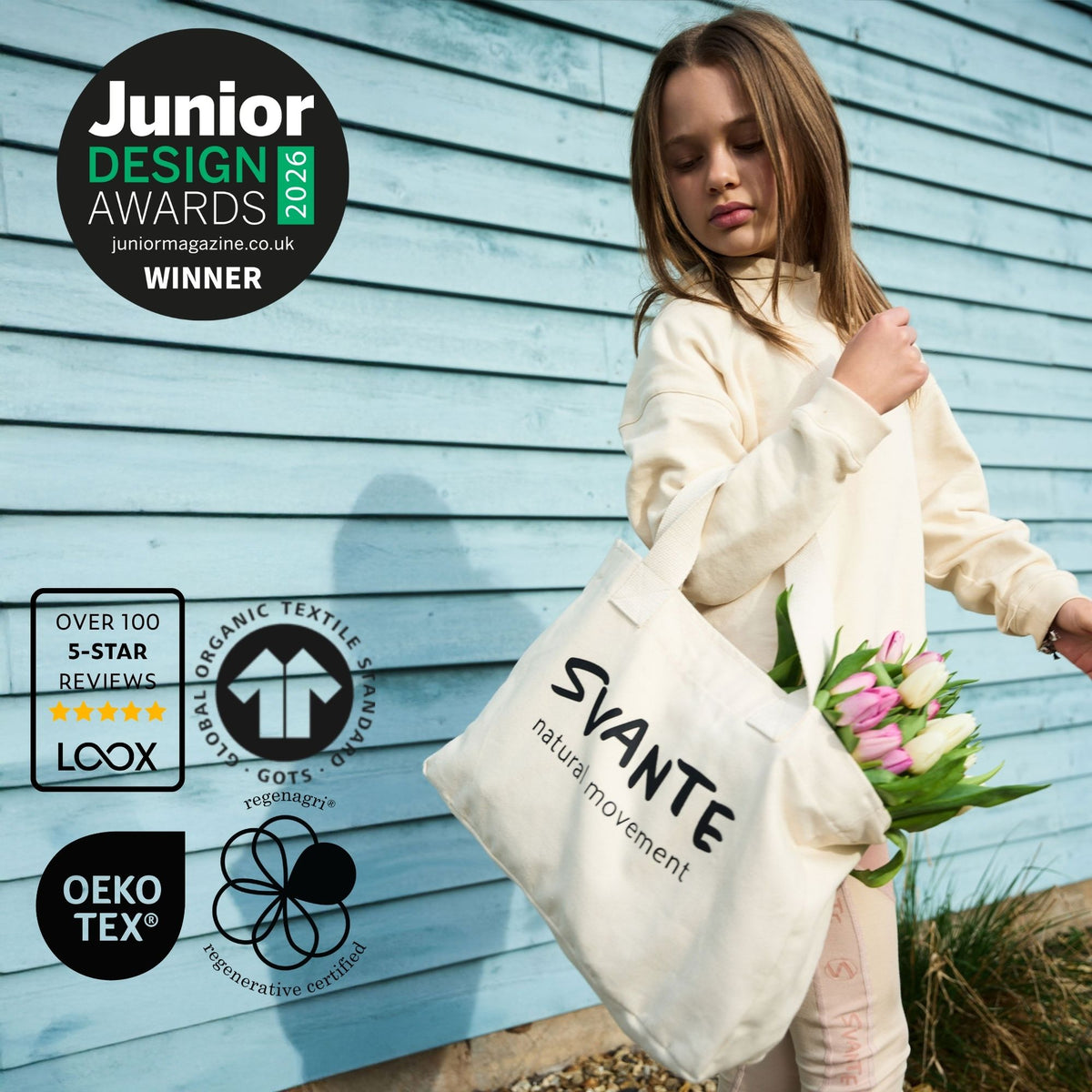 A child wearing a cream Svante sweatshirt holds a cream Svante organic cotton canvas tote bag printed with the brand name 'SVANTE' and 'natural movement' in black text, standing against a light blue wooden wall and holding pink tulips. Multiple certification and award badges are overlaid on the left side of the image.