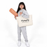 A young girl with long black hair stands against a white studio background, wearing a Svante regenerative cotton light grey hoodie and sweatpants with white sneakers. She is holding a rolled tan exercise mat and carrying a large cream canvas tote bag printed with the brand name 'SVANTE' and the text 'natural movement. 