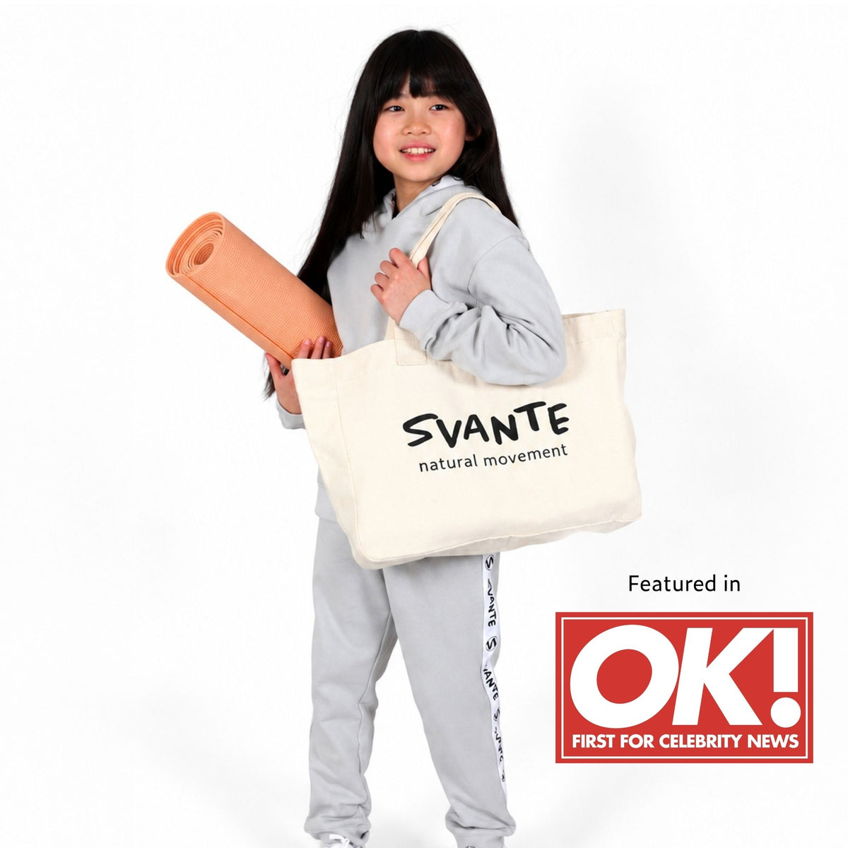 A young girl in a Svante organic cotton light grey tracksuit holds a Svante organic cotton cream canvas tote bag printed with 'SVANTE natural movement' and carries a rolled peach yoga mat. A red 'Featured in OK! FIRST FOR CELEBRITY NEWS' badge is shown in the bottom right corner.