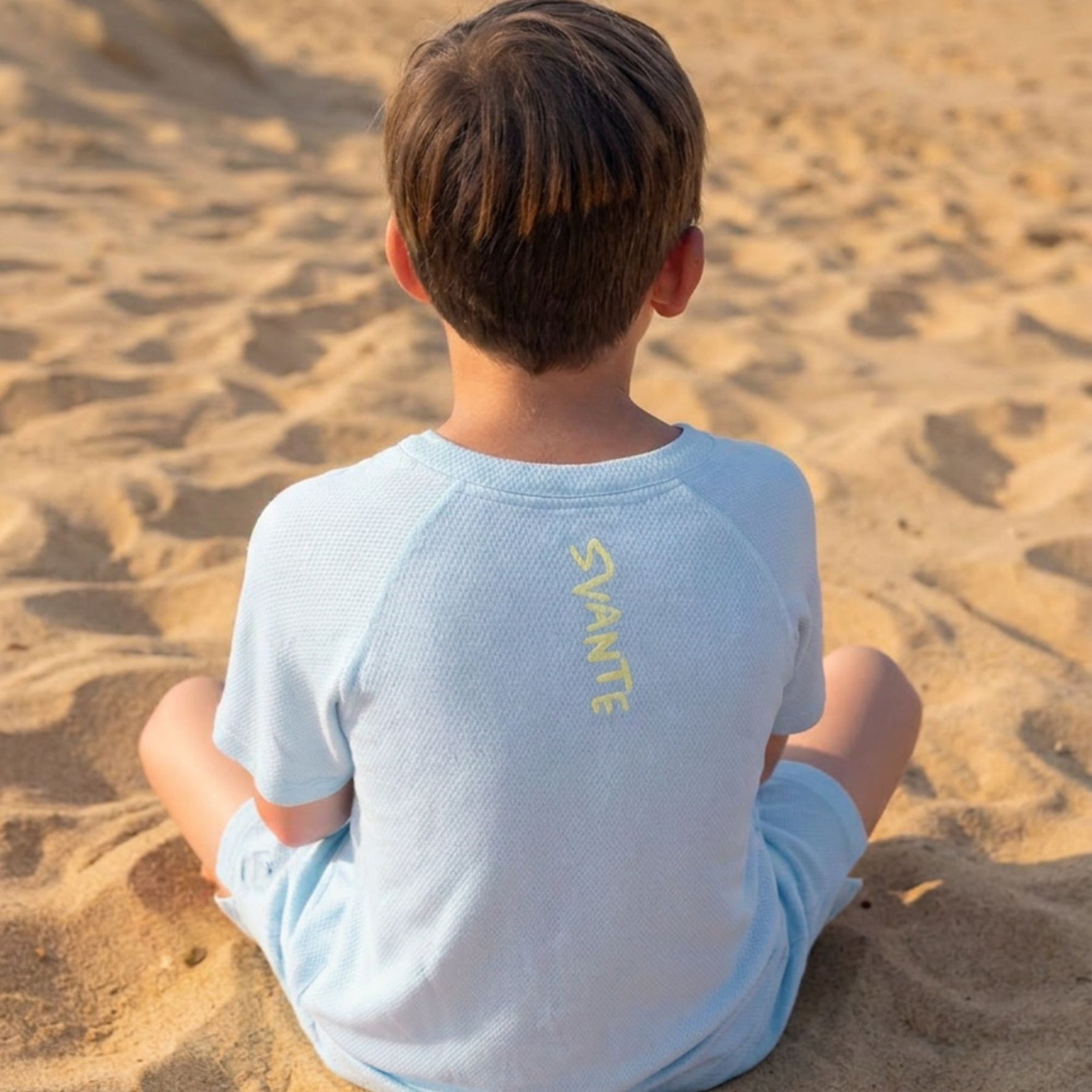 Boy sitting on a sandy beach wearing a light blue all natural plastic free sports t-shirt and short set with 'SVANTE' branding.
