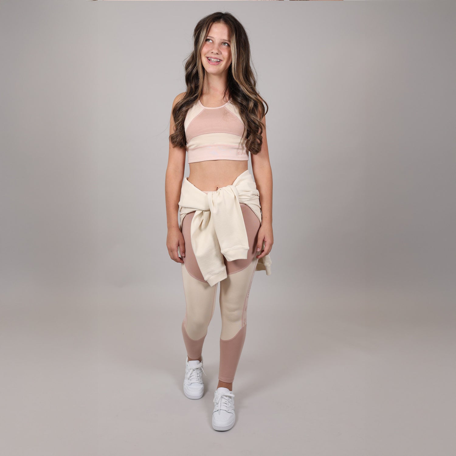 Young girl front facing with big smile and wearing a beige sports crop-top and beige leggings with a matching hoodie tied around her waist on a grey background