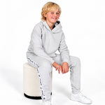 A young blonde boy wearing a Svante  light grey regenerative cotton hooded sweatshirt and matching jogger pants with white sneakers, sitting on a cylindrical cream-colored stool against a white studio background. 