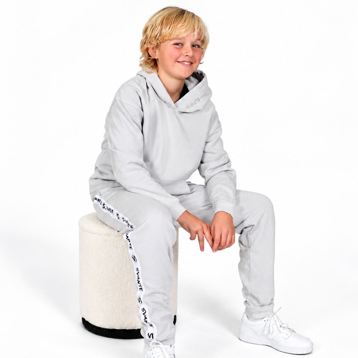A young blonde boy wearing a Svante  light grey regenerative cotton hooded sweatshirt and matching jogger pants with white sneakers, sitting on a cylindrical cream-colored stool against a white studio background. #Harbour-Mist