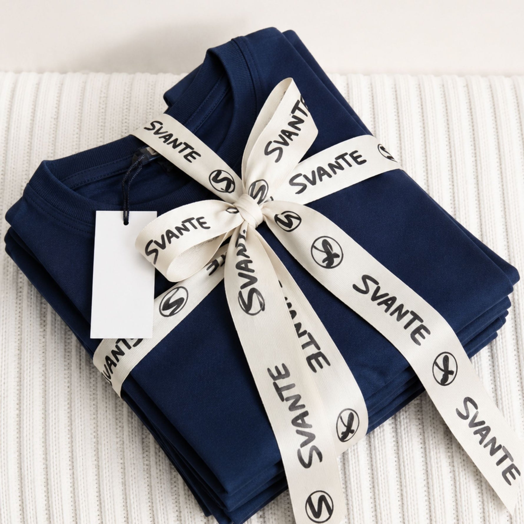 Folded organic cotton navy blue T-shirts with a white ribbon and 'SVANTE' label on a textured surface. 