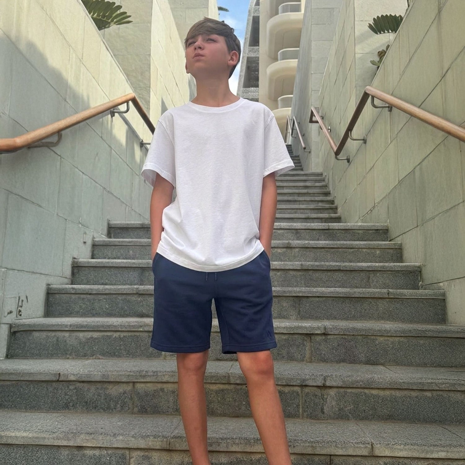 Boy wearing an organic cotton white t-shirt and organic cotton blue jersey shorts standing on a set of stairs. 