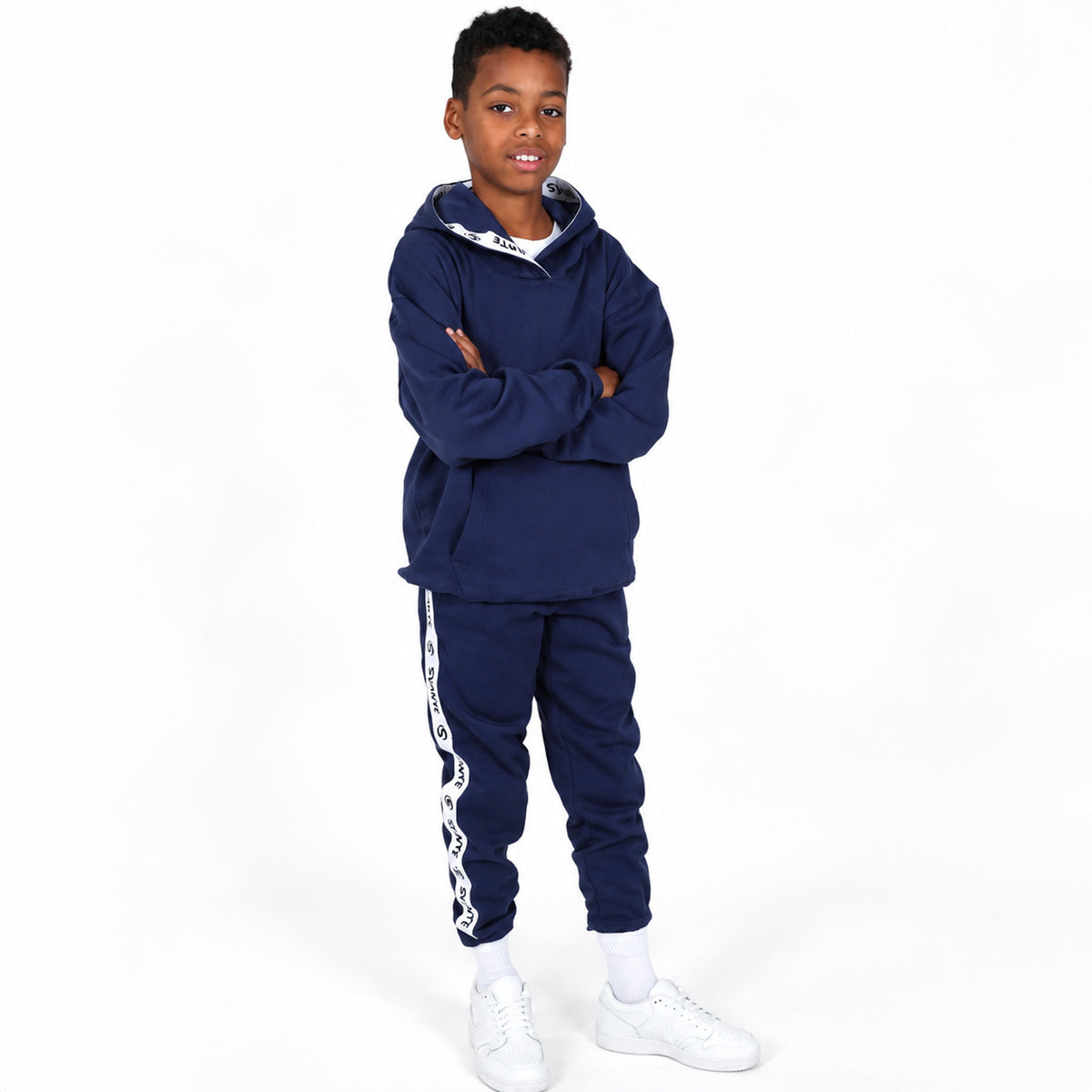 A young boy modelling a navy blue two-piece Svante regenerative cotton tracksuit with white graphic details, consisting of a pullover hoodie and matching jogger pants, paired with white sneakers against a plain white background. 