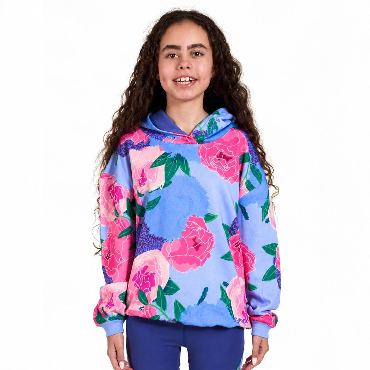 A young girl with long, curly brown hair stands against a solid white background wearing a Svante regenerative cotton light blue hooded sweatshirt with a vibrant pink and green floral print, paired with dark blue pants.