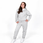 A girl modelling a Svante regenerative cotton light grey two-piece loungewear set consisting of a pullover hoodie and matching sweatpants, paired with white sneakers, against a plain white studio background.
