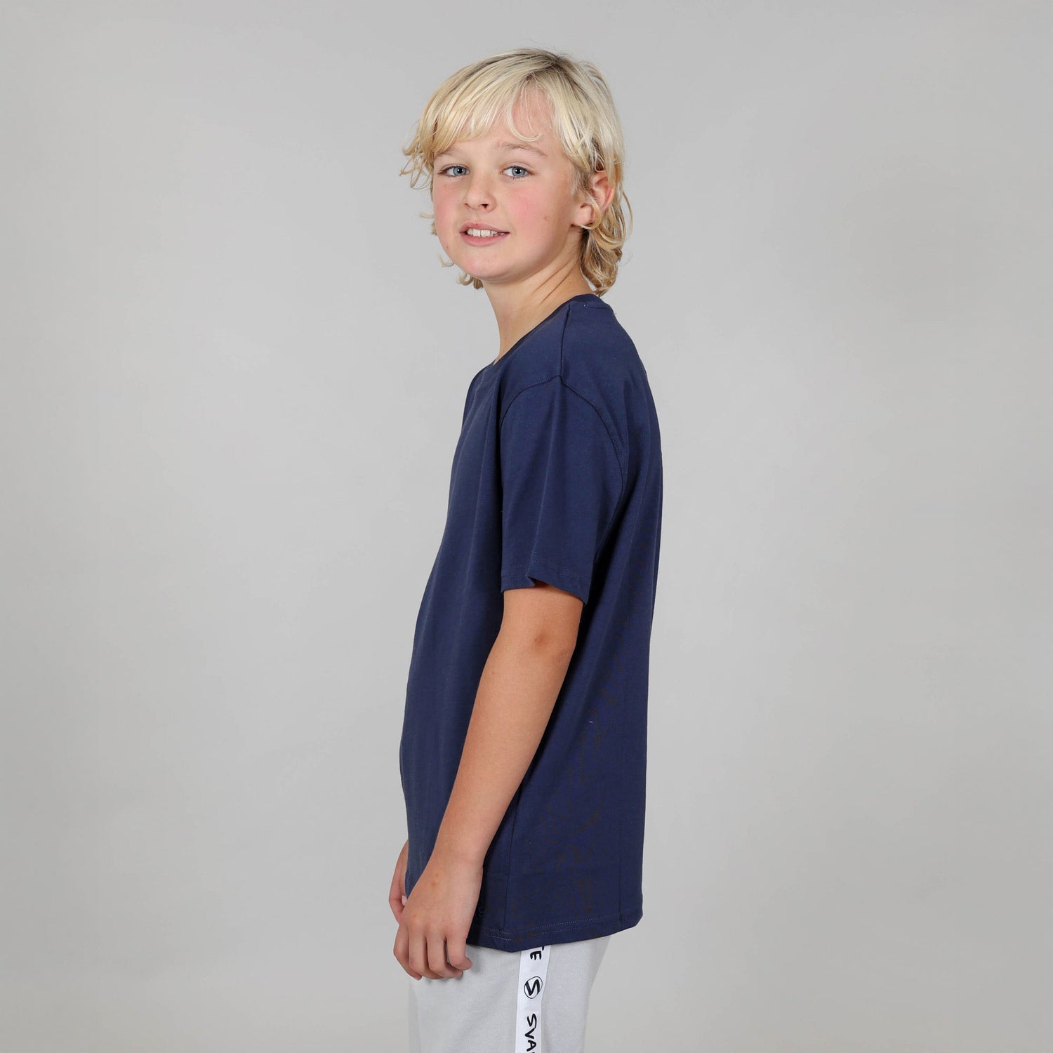 Boy aged ten, standing side on wearing a navy blue organic relaxed fit t-shirt against a grey background. 