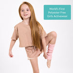 Young girl wearing pink organic cotton crop t shirt and pink sports shorts, with text 'World's First Polyester Free Girls Activewear' on a light blue sign, on white background. 