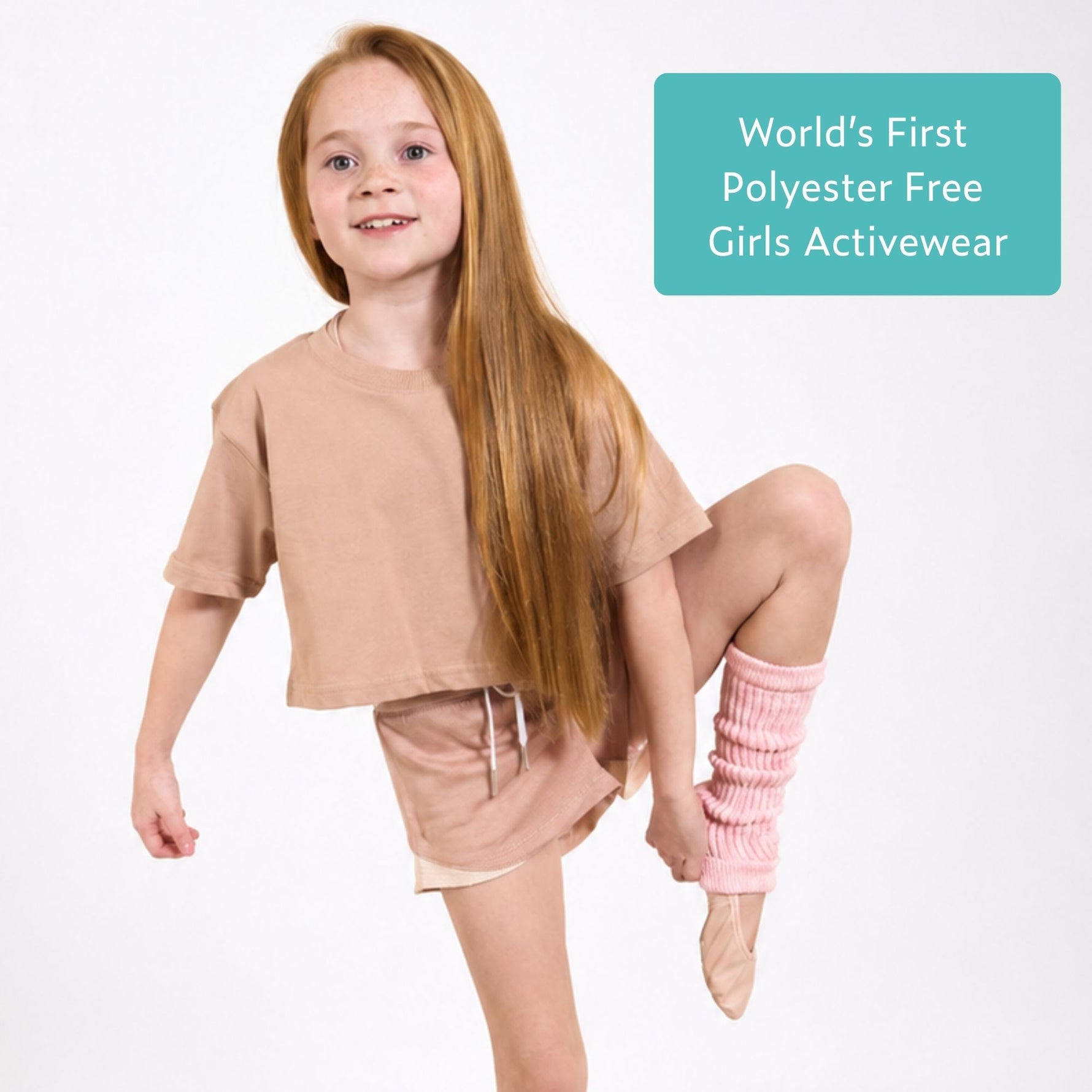 Young girl wearing pink organic cotton crop t shirt and pink sports shorts, with text 'World's First Polyester Free Girls Activewear' on a light blue sign, on white background. 