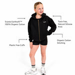 A young girl wearing a Svante organic cotton black zip-up hoodie with a gold zipper and the brand name 'SVANTE' embroidered in white on the chest, paired with black shorts and a black crop top, set against a plain white background. Call out signs for Earthsoft organic cotton and plastic free cuffs. 