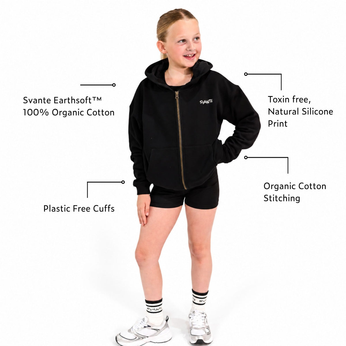 A young girl wearing a Svante organic cotton black zip-up hoodie with a gold zipper and the brand name 'SVANTE' embroidered in white on the chest, paired with black shorts and a black crop top, set against a plain white background. Call out signs for Earthsoft organic cotton and plastic free cuffs. 