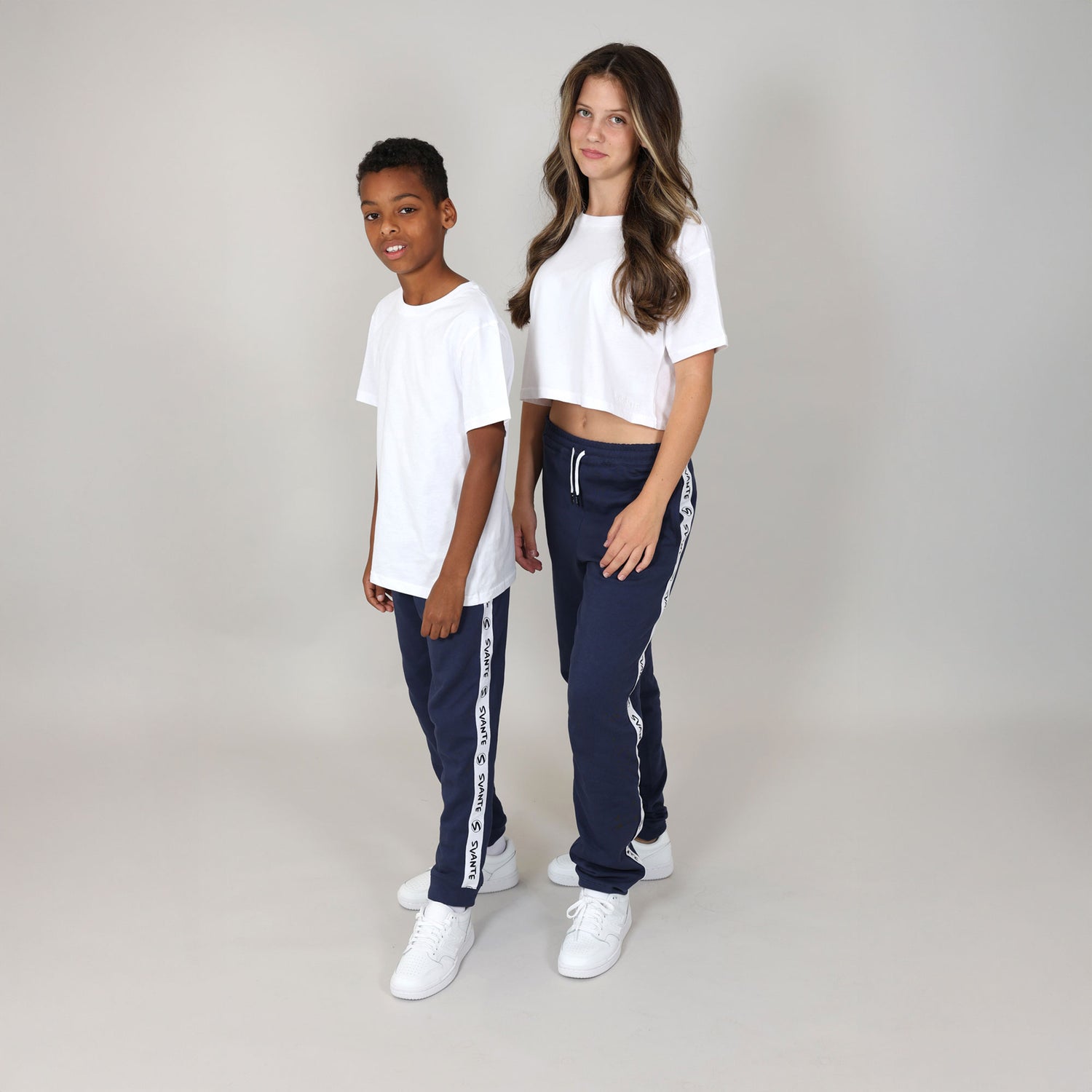 Boy and girl wearing matching unisex white organic t-shirts and navy tracksuits with white logo stripe down leg on a plain background