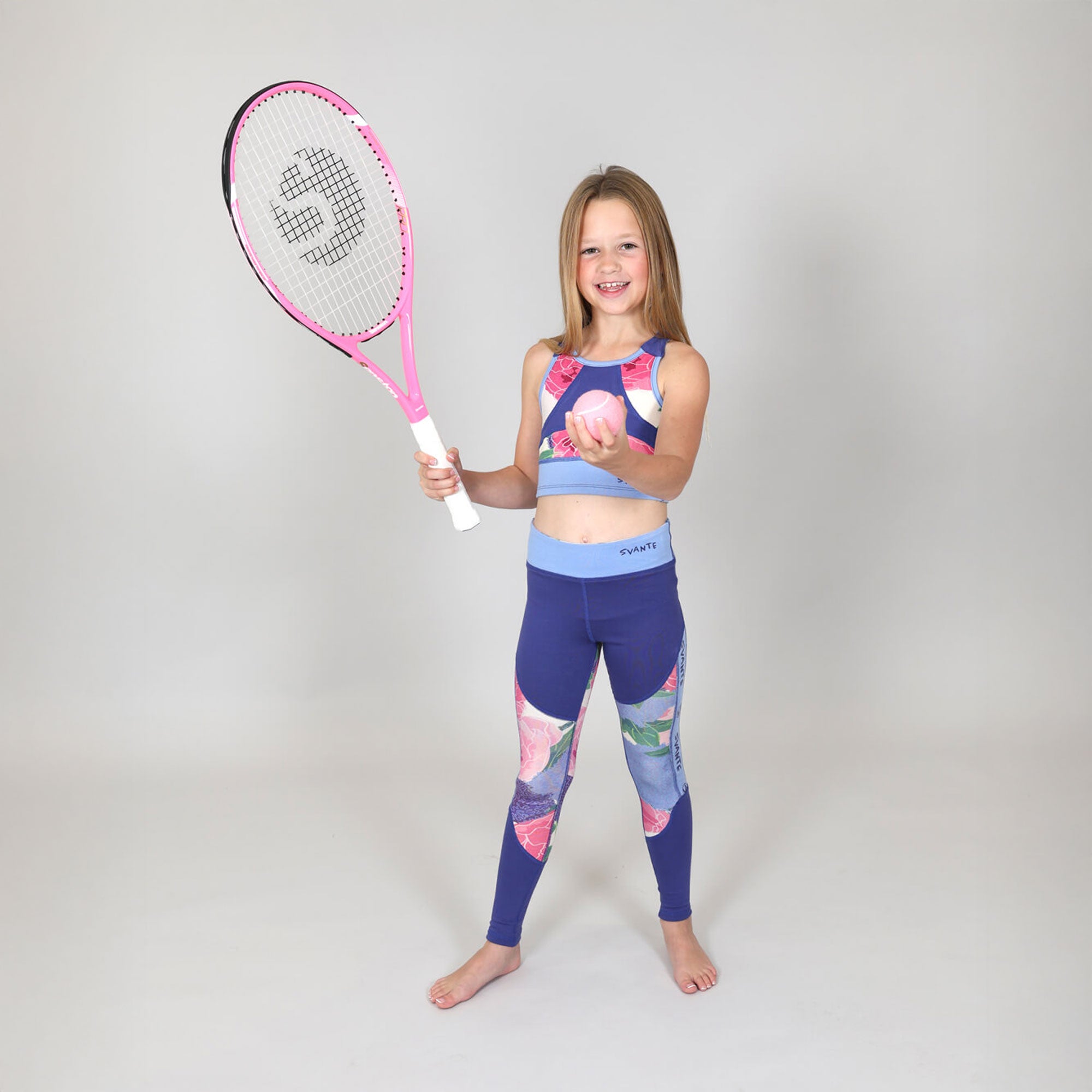 Young girl in sports crop-top and matching leggings holding a pink tennis racket against a plain background