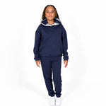 A girl model wearing a Svante regenerative cotton navy blue sweatshirt and matching sweatpants set, with a white Svante patterned hood lining, standing against a plain white studio background.