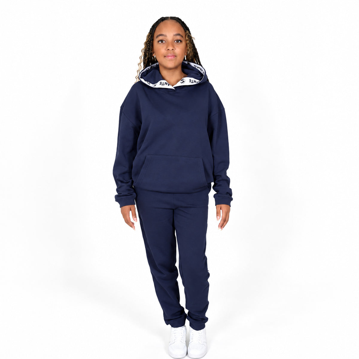 A girl model wearing a Svante regenerative cotton navy blue sweatshirt and matching sweatpants set, with a white Svante patterned hood lining, standing against a plain white studio background.