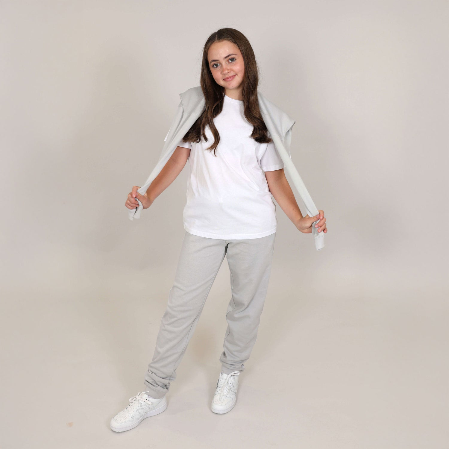 Teenage girl with nice smile wearing organic grey joggers, with white t-shirt and white trainers, she has hoodie over her shoulders and pulling the arms out to the side. 