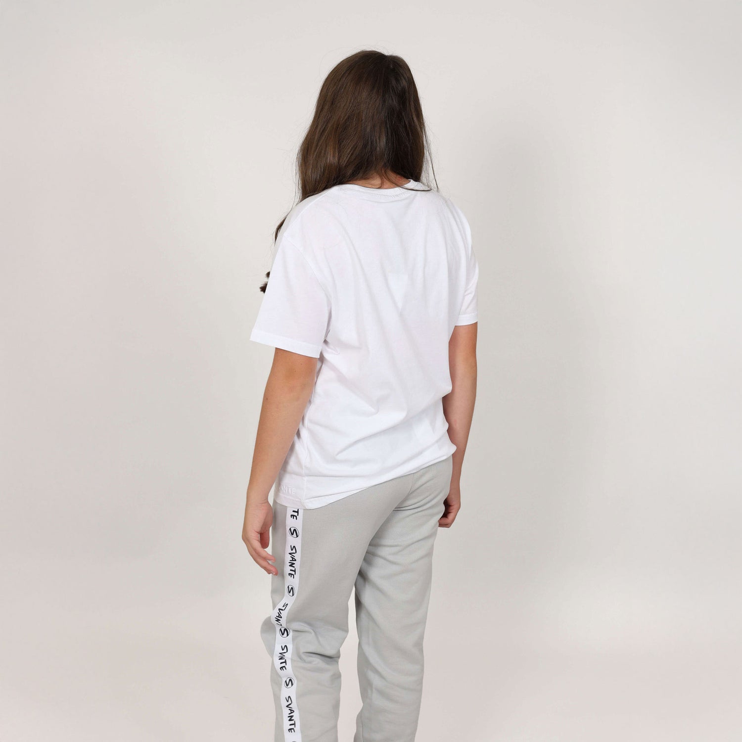 Girl facing away from camera wearing a white organic t-shirt and light grey joggers  with 'Svante' branding down the side on a plain background. 