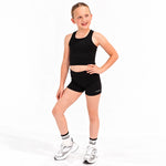 A young girl modelling a Svante black athletic crop top and matching black sports  shorts, paired with white and silver sneakers and white socks, against a plain white studio background.
