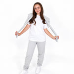 A young girl modeling a white Svante organic cotton crewneck t-shirt, and Svante regenerative cotton  light grey sweatpants, and white sneakers, holding a light gray zip-up hoodie open over her shoulders against a white studio background. 