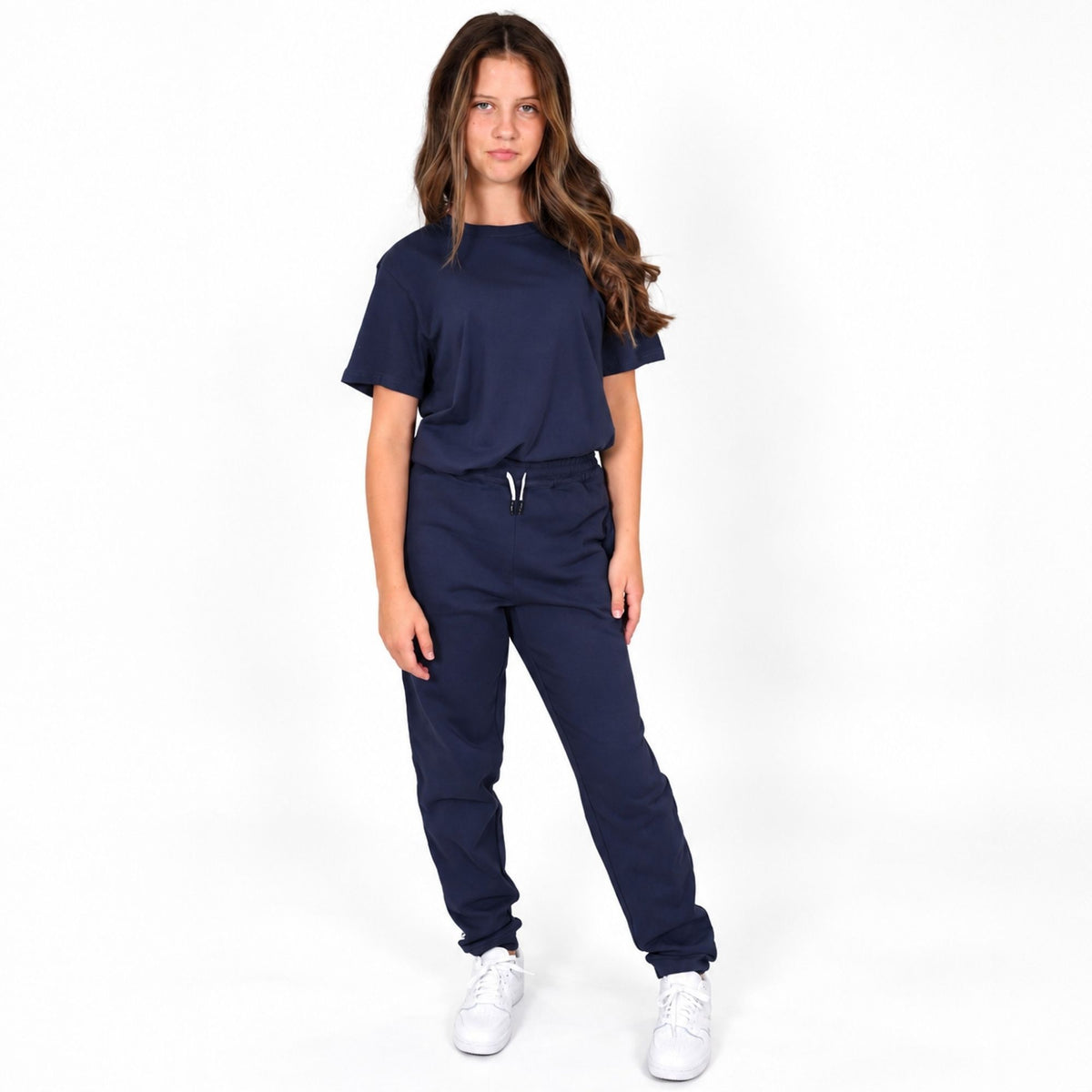 A young girl modelling a matching Svante regenerative cotton navy blue joggers, consisting of an organic cotton short-sleeve crewneck t-shirt and jogger-style pants with a drawstring waist, paired with white sneakers, against a plain white studio background. 