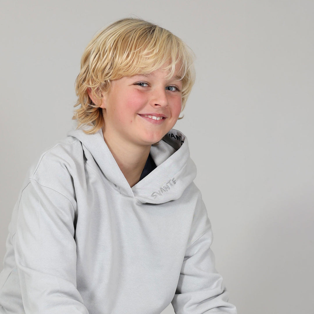 Boy smiling wearing a light grey hoodie with 'Svante' logo a plain neutral background. 